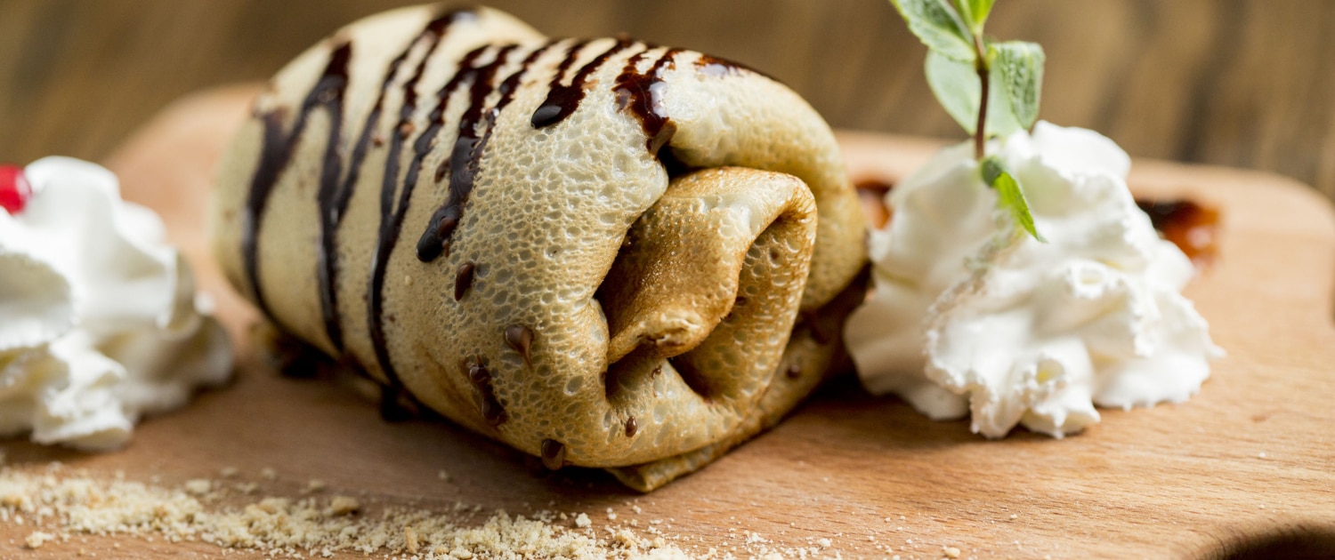 Sweet crepe rolled up and drizzled with chocolate beside piped cream