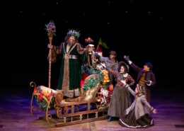 A group of townspeople surround the Ghost of Christmas Present on his sleigh