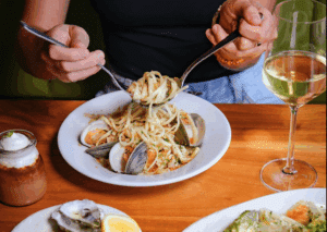 A woman twirling pasta onto a fork with wine and oysters on the table
