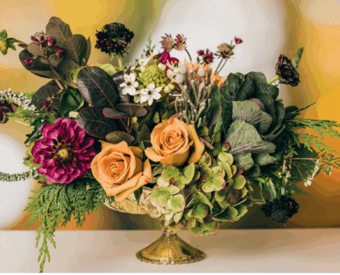 A floral arrangement with greenery, muted yellow tones, and a deep red flower