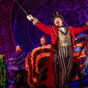 A performer with goatee, red coat, gold vest, and tophat in front of Can-Can dancers at the Moulin Rouge