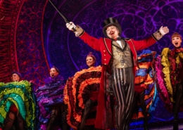 A performer with goatee, red coat, gold vest, and tophat in front of Can-Can dancers at the Moulin Rouge