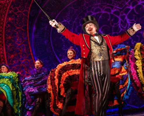 A performer with goatee, red coat, gold vest, and tophat in front of Can-Can dancers at the Moulin Rouge