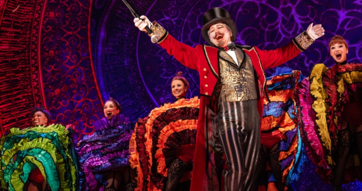 A performer with goatee, red coat, gold vest, and tophat in front of Can-Can dancers at the Moulin Rouge