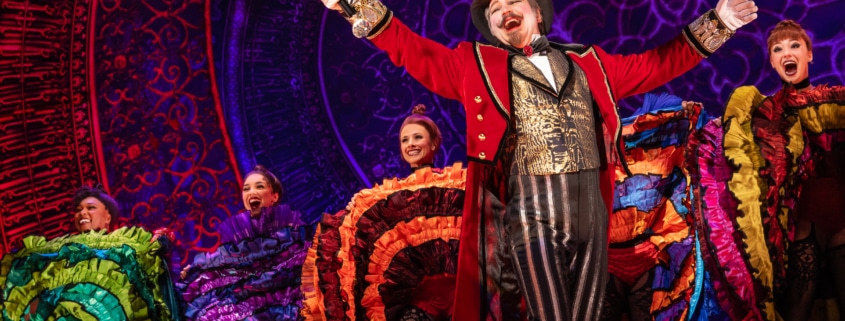 A performer with goatee, red coat, gold vest, and tophat in front of Can-Can dancers at the Moulin Rouge