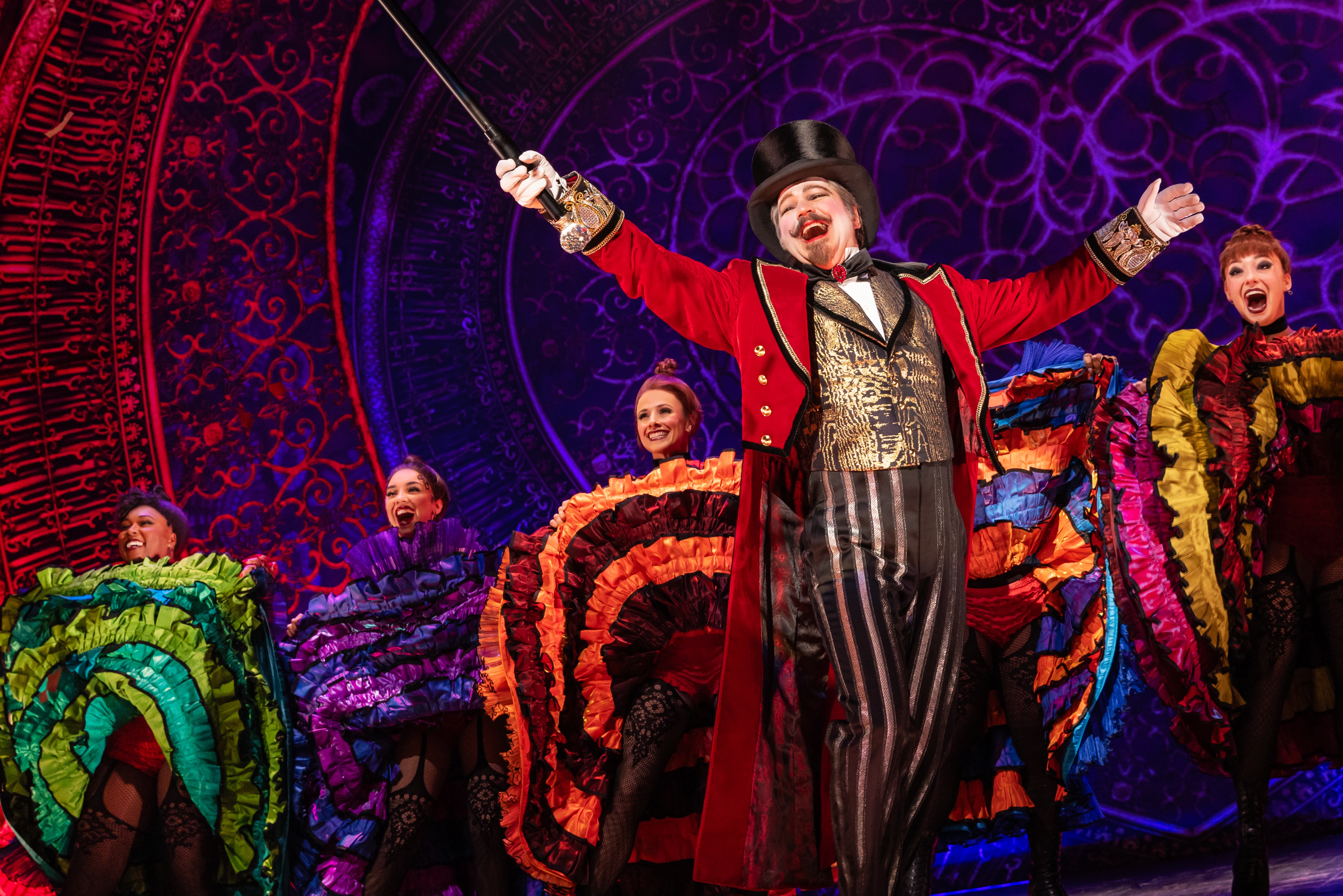 A performer with goatee, red coat, gold vest, and tophat in front of Can-Can dancers at the Moulin Rouge