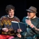 A young man in a colorful sweater sits on a park bench next to his grandfather who is wearing a green track suit and a fedora. They are laughing and reading a book together