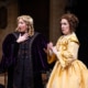 A young girl with curly blonde hair, wearing an intricate purple velvet robe and an older woman, her mother, in a bright yellow frock look concerned.