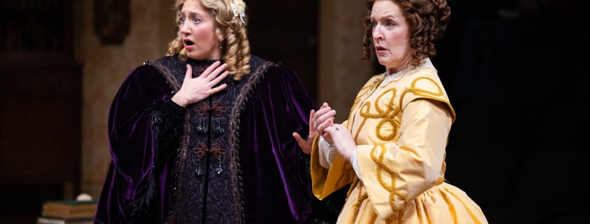 A young girl with curly blonde hair, wearing an intricate purple velvet robe and an older woman, her mother, in a bright yellow frock look concerned.