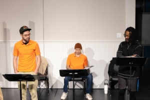 Three actors rehearse at music stands. Two are standing and one is seated.