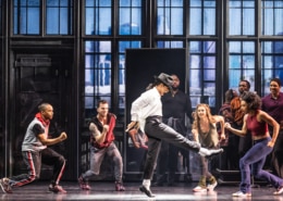 Michael Jackson dances in rehearsal, wearing the signature black and white ensemble. Backup dancers join in, facing him.