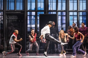 Michael Jackson dances in rehearsal, wearing the signature black and white ensemble. Backup dancers join in, facing him.