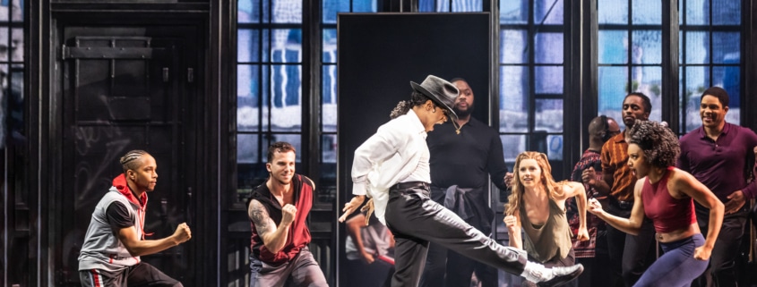 Michael Jackson dances in rehearsal, wearing the signature black and white ensemble. Backup dancers join in, facing him.