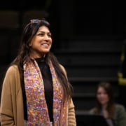 An actor stands onstage, smiling, during rehearsal. She is wearing a colorful scarf and a tan sweater.