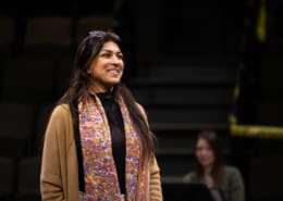 An actor stands onstage, smiling, during rehearsal. She is wearing a colorful scarf and a tan sweater.