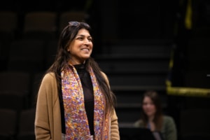 An actor stands onstage, smiling, during rehearsal. She is wearing a colorful scarf and a tan sweater.