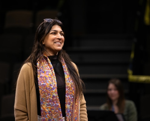 An actor stands onstage, smiling, during rehearsal. She is wearing a colorful scarf and a tan sweater.