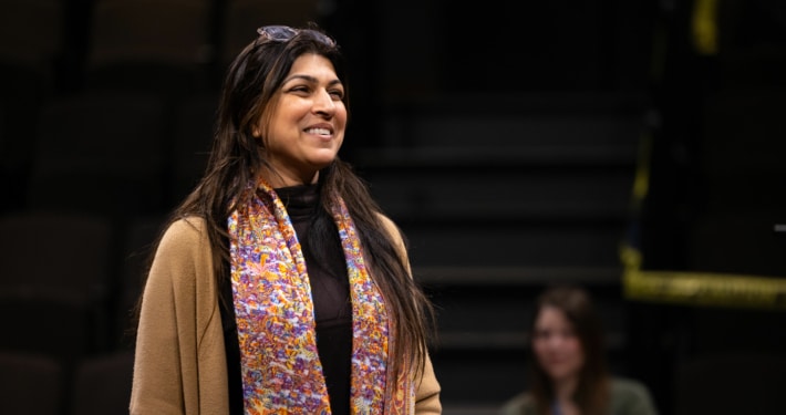An actor stands onstage, smiling, during rehearsal. She is wearing a colorful scarf and a tan sweater.