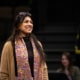 An actor stands onstage, smiling, during rehearsal. She is wearing a colorful scarf and a tan sweater.