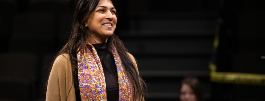 An actor stands onstage, smiling, during rehearsal. She is wearing a colorful scarf and a tan sweater.