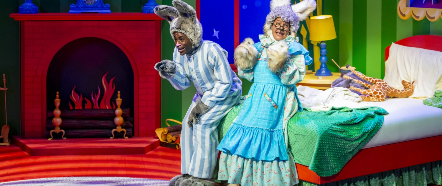 A photo of two actors wearing rabbit ears and laughing. The set is designed in primary colors with a fireplace, bed, lamp, curtains, and rug.