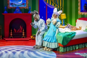 A photo of two actors wearing rabbit ears and laughing. The set is designed in primary colors with a fireplace, bed, lamp, curtains, and rug.