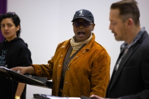 A closeup shot of three actors at their music stands. They are looking at one another as they rehearse.