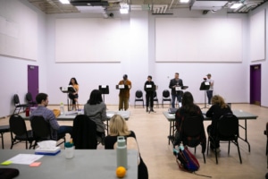 A group of actors stand in a row at music stands. The creative team sits at tables facing them, away from the camera.