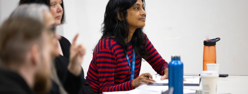 Playwright Vauhini Vara sits at a table with her peers. Papers and drinks are scattered across the table, the team is hard at work.