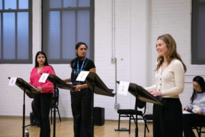 Three women stand at music stands in a rehearsal room. Two are looking at the other, concerned, while the third has a smile on her face.