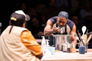 A man in a orange shirt and trucker hat watches another Black man in a muscle shirt carefully season a pot.