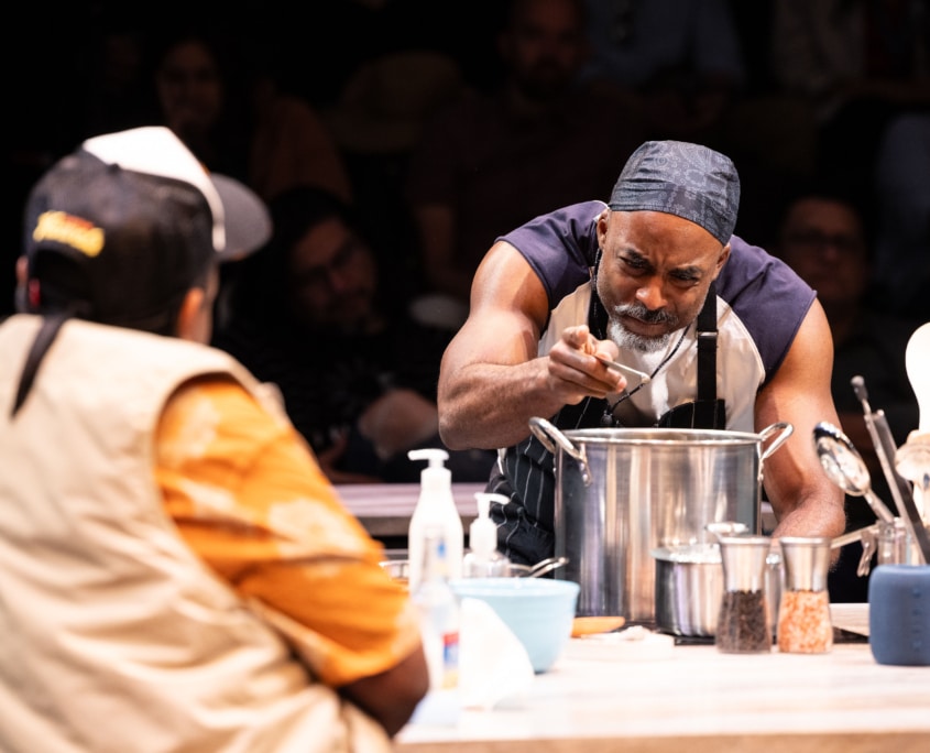 A man in a orange shirt and trucker hat watches another Black man in a muscle shirt carefully season a pot.