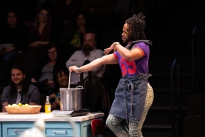 A Black man with beaded braids, wearing a purple and red jersey and a half apron stirs a stainless-steel pot.