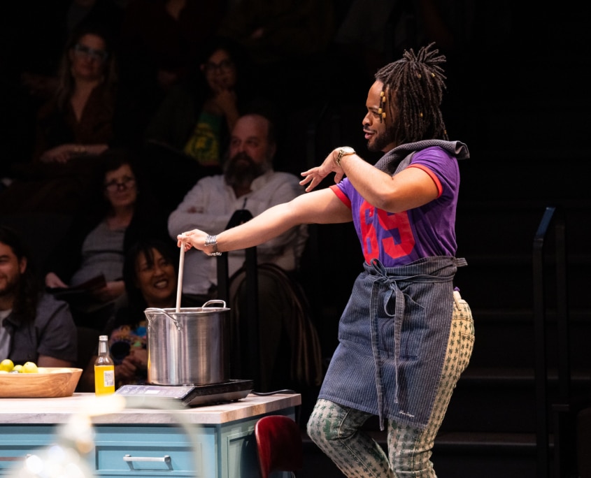 A Black man with beaded braids, wearing a purple and red jersey and a half apron stirs a stainless-steel pot.