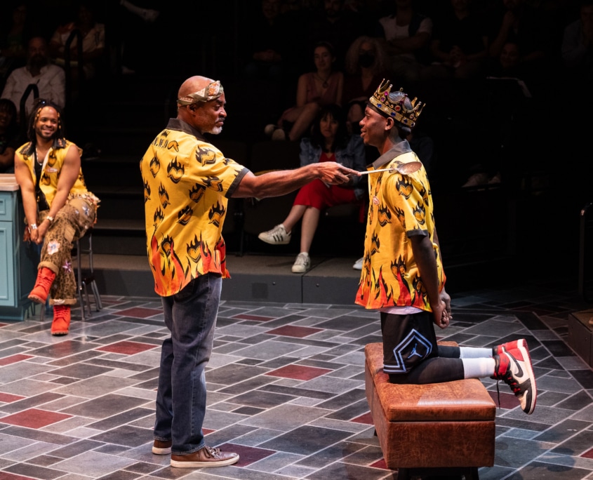 An older Black man knights a younger Black man with a large, slotted spoon, another Black man watches the scene, smiling. The young man is wearing a gold crown, all the men are wearing yellow and orange Hawiian shirts with flaming hearts on them.