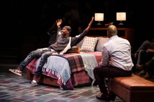A young Black man in a black windbreaker and ripped jeans leans back on a neatly made bed with, smiling with his arms spread wide. A Black man in a neat button-down shirt and dress shoes faces him.