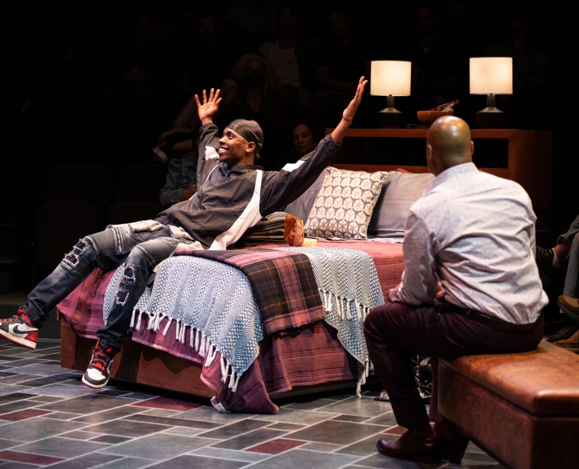 A young Black man in a black windbreaker and ripped jeans leans back on a neatly made bed with, smiling with his arms spread wide. A Black man in a neat button-down shirt and dress shoes faces him.