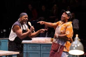 Two Black men in casual modern clothing and aprons sing into a wooden spoon in a kitchen.