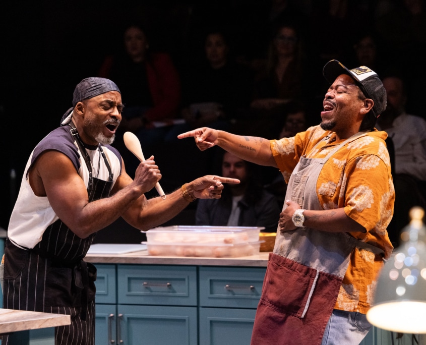 Two Black men in casual modern clothing and aprons sing into a wooden spoon in a kitchen.