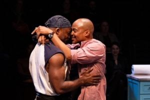 Two Black men, one wearing a muscle shirt and an apron, the other wearing a pink button down and loafers, embrace one another tightly.