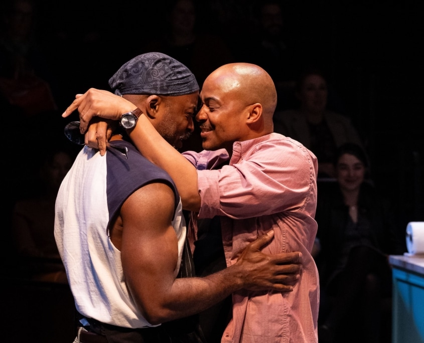 Two Black men, one wearing a muscle shirt and an apron, the other wearing a pink button down and loafers, embrace one another tightly.