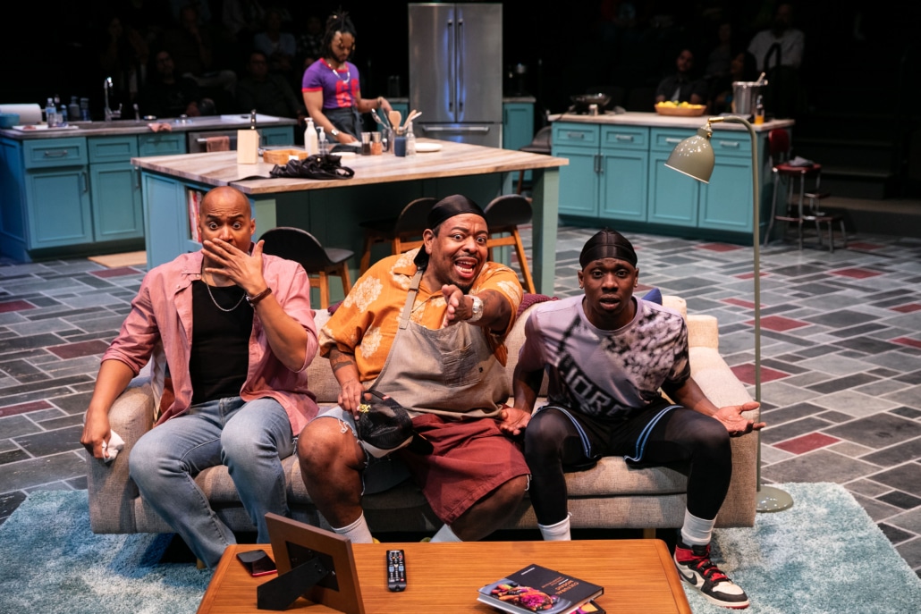 Three Black men sit on the couch they are watching TV and reacting to what is happening on the screen, a fourth Black man stands at a kitchen island behind them.