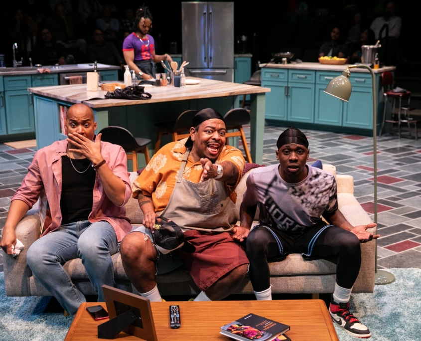 Three Black men sit on the couch they are watching TV and reacting to what is happening on the screen, a fourth Black man stands at a kitchen island behind them.