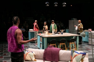 Four Black men in a kitchen look back confused at a fifth Black man who is facing away from the camera, wearing a purple sleeveless shirt.