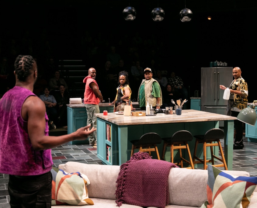 Four Black men in a kitchen look back confused at a fifth Black man who is facing away from the camera, wearing a purple sleeveless shirt.