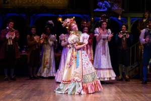 Stephanie Lynne Mason. Photo by Jamie Kraus Photography. Alt Text: Mrs. Fezziwig stands center stage in an elaborate, colorful Victorian gown adorned with floral accents and ribbons, holding a fan and striking a pose.