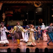 A joyous dance scene with performers in vibrant Victorian costumes holding hands and moving energetically across the stage. The background features holiday decorations, including a Christmas tree and ornate signage, adding to the celebratory mood.