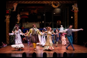 Vi Van Dang, Shabazz Green, Stephanie Lynne Mason, and Cordell Cole. Photo by Jamie Kraus Photography. A joyous dance scene with performers in vibrant Victorian costumes holding hands and moving energetically across the stage. The background features holiday decorations, including a Christmas tree and ornate signage, adding to the celebratory mood.