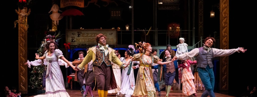 A joyous dance scene with performers in vibrant Victorian costumes holding hands and moving energetically across the stage. The background features holiday decorations, including a Christmas tree and ornate signage, adding to the celebratory mood.