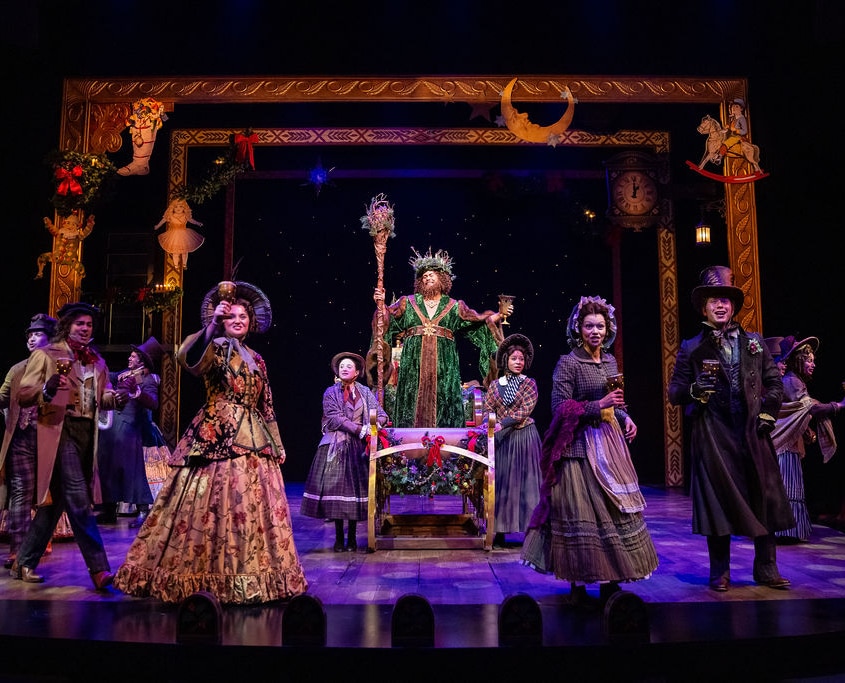 Topher Embrey and the cast of A Christmas Carol. Photo by Jamie Kraus Photography. The Ghost of Christmas Present stands on his sleigh dressed in a grand green robe and crown, holding a staff. The ensemble dressed in ornate period costumes gathered around him.